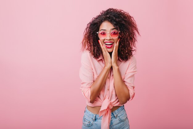 Tolles afrikanisches Modell, das beim Posieren überraschte Gefühle ausdrückt. Stilvolle lockige Frau in der Sonnenbrille, die Spaß hat.