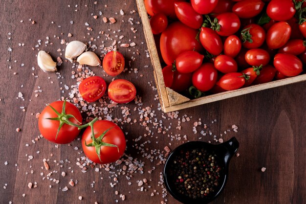 Tomaten in der Holzkiste nahe dem Pulver des schwarzen Pfeffers im schwarzen Schüsselknoblauch und Tomaten verbreiteten Seesalz auf brauner Steinoberflächenhahnansicht