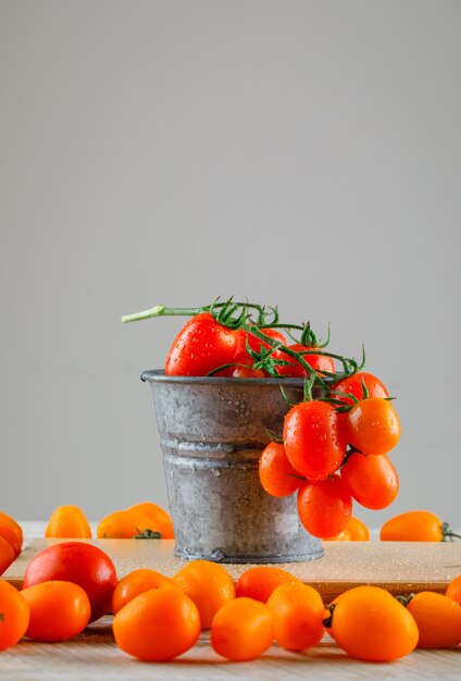 Tomaten in einem Mini-Eimer mit Schneidebrett-Seitenansicht auf hölzernem und grauem Tisch