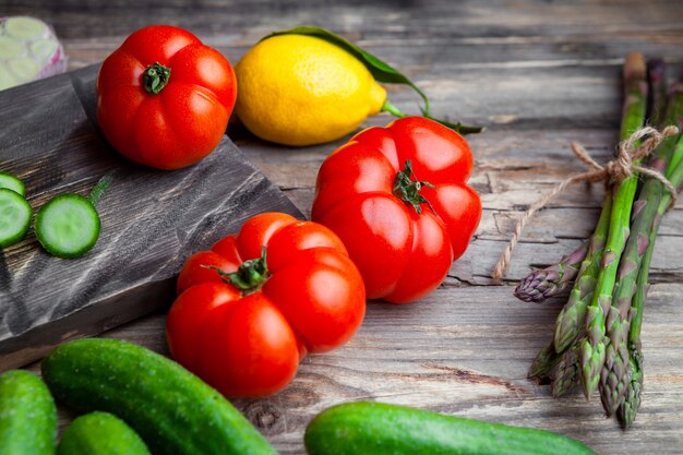 Tomaten mit hohem Blickwinkel in Schneidebrett mit geschnittener Gurke, Knoblauch, Spargel, Zitrone auf dunklem hölzernem Hintergrund. horizontal