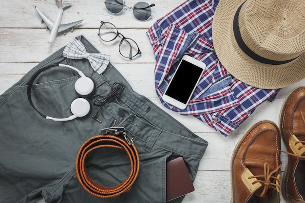 Top-Ansicht-Accessoires, um mit Mann-Kleidung-Konzept zu reisen. Hemd, Jeans, Handy, Kopfhörer auf Holzuntergrund.passport, Schlüssel, Brillen und Schuhe auf Holztisch.