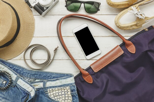 Top-Ansicht Zubehör zu reisen mit Frauen Kleidung concept.white Mobiltelefon, Gürtel, Tasche, Hut, Kamera, Halskette, Hose und Sonnenbrille auf weißem Holz Tisch.