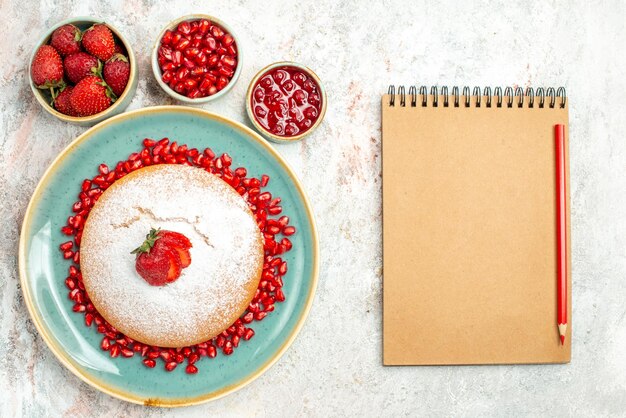 Top Nahaufnahme appetitlicher Kuchen ein appetitlicher Kuchen mit Granatapfel neben den Schalen mit Erdbeeren und Granatapfel und Sahne-Notizbuch mit Rotstift auf dem Tisch