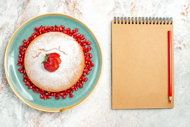 Top Nahaufnahme Erdbeerkuchen Creme Notizbuch Rotstift neben dem Kuchenteller mit Erdbeeren und Granatapfel auf dem Tisch