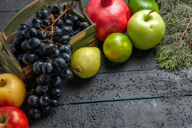Top Nahaufnahme Früchte Trauben in Holzkiste Äpfel Granatapfel Birnen Limetten neben Fichtenzweigen auf dunklem Tisch