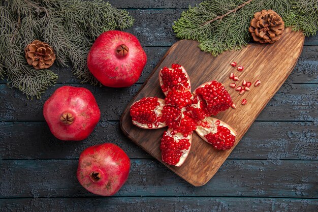 Top Nahaufnahme Granatapfel und Zweige pilled Granatapfel auf Holzbrett neben Samen von Granatapfel Fichtenzweigen mit Kegelmesser und drei Granatäpfel auf grauem Tisch