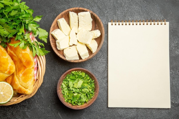 Top Nahaufnahme Käsekräuter weißes Notizbuch neben den Schüsseln mit Kräutern und Käse und Korb mit appetitlichen Kuchen Zitronenkräuter und karierte Tischdecke