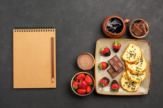 Top-Nahaufnahme-Kuchen mit Schokoladennotizbuch und Bleistift neben dem Teller mit appetitlichem Kuchen mit Schokolade und Erdbeeren und Erdbeeren und Schokoladencreme in Schalen