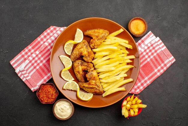 Top Nahaufnahme leckere Hähnchen appetitlich Chicken Wings Pommes frites und Zitronenschalen mit verschiedenen Saucen und Gewürzen auf rosa-weiß karierter Tischdecke in der Mitte des dunklen Tisches