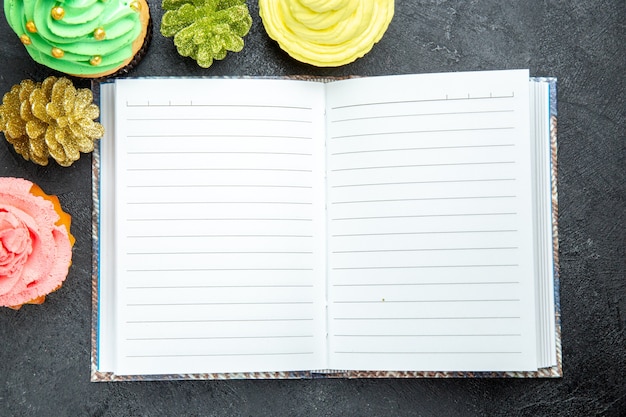 Top Nahaufnahme Mini bunte Cupcakes und ein Notizbuch auf dunklem Hintergrund