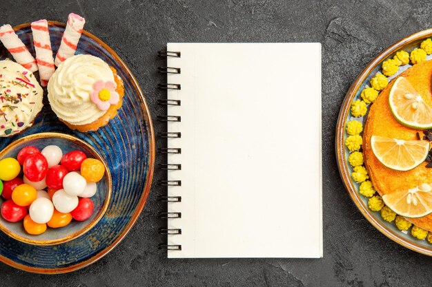 Top Nahaufnahme Süßigkeiten auf dem Teller appetitliche Cupcakes und Schüssel mit Süßigkeiten neben dem weißen Notebook-Teller mit Kuchen mit Zitrusfrüchten auf dem schwarzen Tisch