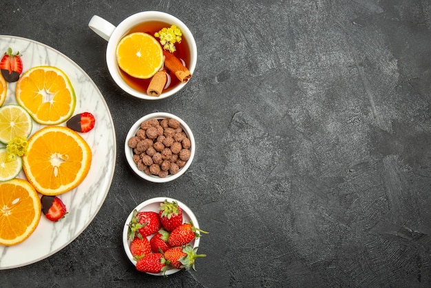 Top Nahaufnahme Tasse Tee und Früchte eine Tasse Tee mit Zitronen- und Zimtschalen mit Nüssen und einem Teller mit Zitrusfrüchten und schokoladenüberzogenen Erdbeeren auf der linken Seite des dunklen Tisches