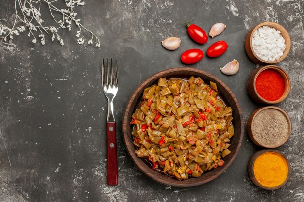 Top Nahaufnahme Teller und Gewürze Teller mit grünen Bohnen und Tomaten Gabel vier Schüsseln mit bunten Gewürzen und Knoblauch auf dem schwarzen Tisch