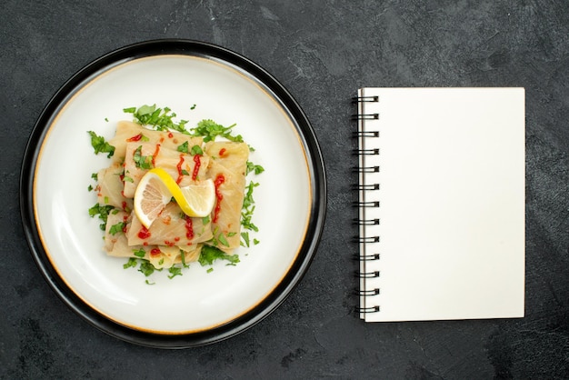 Top-nahaufnahmeplatte mit appetitlichem gericht gefülltem kohl mit kräutern, zitrone und soße auf einem weißen teller neben einem weißen notizbuch auf schwarzer oberfläche