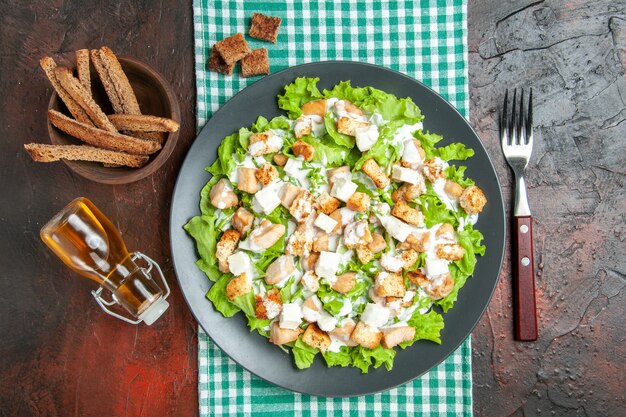 Top View Caesar Salad auf ovaler Platte grün weiß karierte Tischdecke Flaschengabel auf dunkelrotem Hintergrund