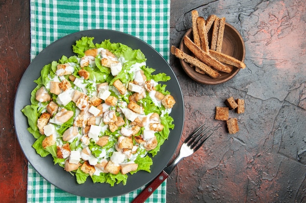 Top View Caesar Salad auf ovaler Platte grün weiß karierte Tischdecke Gabel Schüssel mit getrockneter Kruste auf dunkelrotem Hintergrund