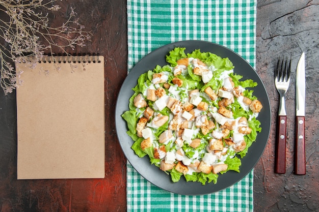 Top view caesar salad auf ovaler platte grün weiß karierte tischdecke notebook messergabel auf dunkelrotem hintergrund Kostenlose Fotos