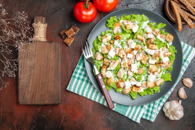 Kostenloses Foto top view caesar salad auf ovaler platte grün-weiß karierten servietten-schneidebrett knoblauch auf dunkelrotem hintergrund