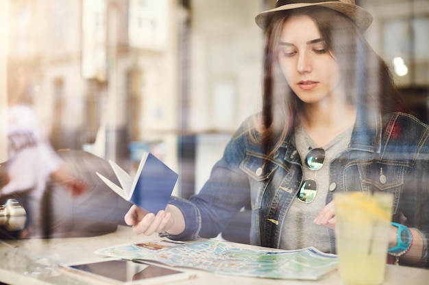 Tourist sitzt im Café und liest eine Karte mit Reisepass, die Limonade in einer geschäftigen Stadt trinkt. Durch Glas geschossen