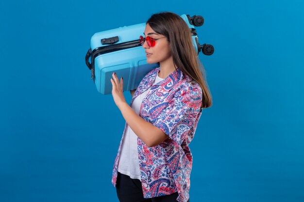 Touristenfrau, die rote Sonnenbrille hält, die Reisekoffer hält, der zuversichtlich schaut, bereit, seitlich stehend auf isoliertem Blau zu reisen