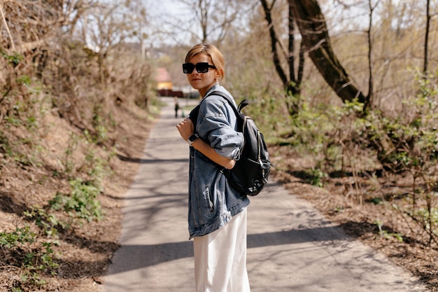 Touristenfrau mit gesammelten hellen Haaren trägt eine Jeansjacke mit Rucksack und dreht sich beim Gehen auf dem Waldweg in die Kamera