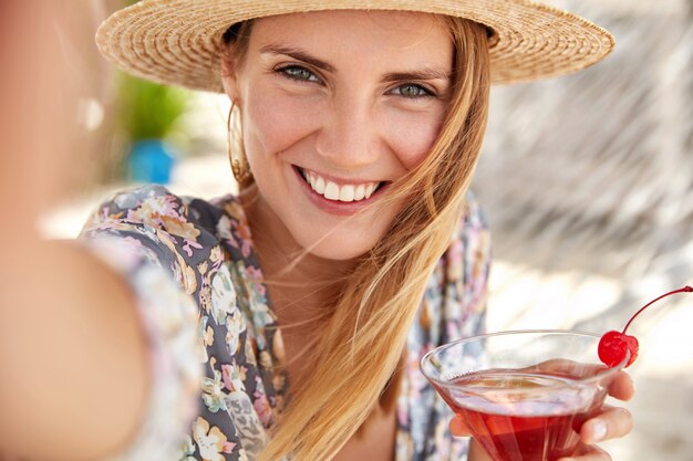 Touristin genießt Sommerferien, trinkt leckeren kalten Cocktail mit Kirsche dekoriert, macht Foto von sich selbst oder Selfie mit nicht erkennbarem Gerät. Sommertourismus, Lifestyle und Ruhekonzept