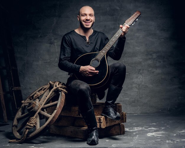 Traditioneller Volksmusiker mit rasiertem Kopf, gekleidet in alte keltische Kleidung, sitzt auf einer Holzkiste und spielt Mandoline.