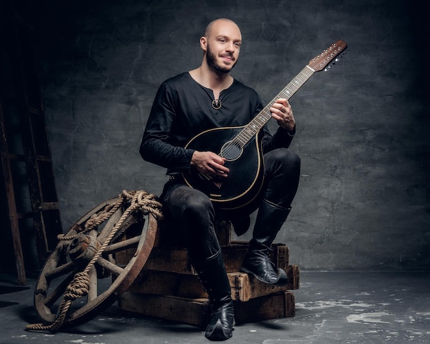 Traditioneller Volksmusiker mit rasiertem Kopf, gekleidet in alte keltische Kleidung, sitzt auf einer Holzkiste und spielt Mandoline.