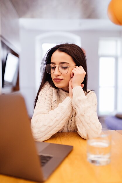 Träumende Dame im weißen Pullover sitzt in der Küche und arbeitet an ihrem Laptop