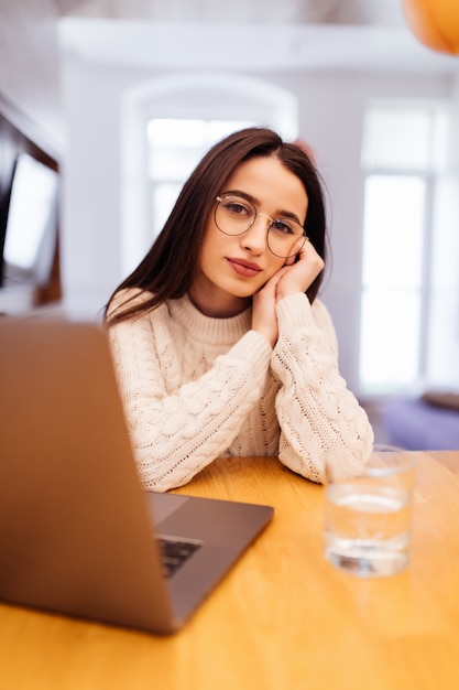 Träumende Dame im weißen Pullover sitzt in der Küche und arbeitet an ihrem Laptop