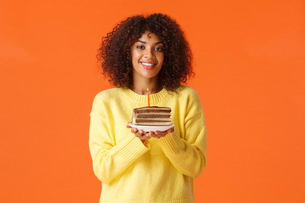 Träumerisches afroamerikanisches b-Tagesmädchen des Taillenporträts mit Afro-Haarschnitt, Teller mit Geburtstagstorte und brennender Kerze haltend, um Wunsch zu machen, glücklich lächelnd, über orange Wand feierend.