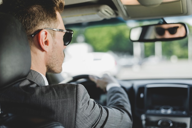 Tragende Sonnenbrille des gutaussehenden Mannes, die Rückspiegel im Auto betrachtet