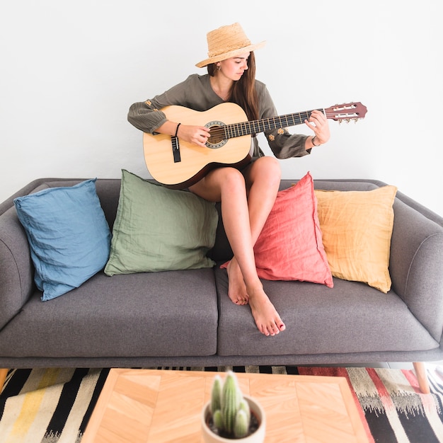 Kostenloses Foto tragender hut der schönen jugendlichen, der auf dem sofa spielt gitarre sitzt