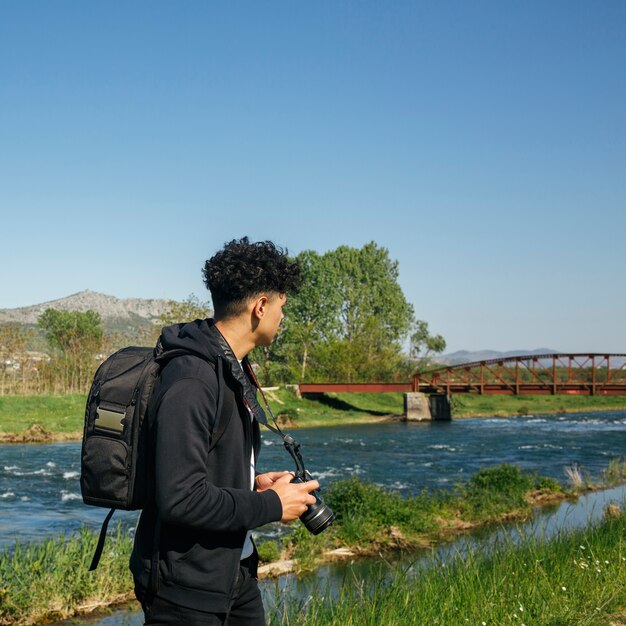 Tragender Rucksack und Kamera des männlichen Fotografen, die nahe schönem Fluss wandern