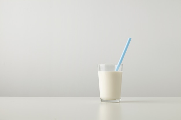 Transparentes Glas mit frischer Bio-Milch und blauem Trinkhalm innen lokalisiert auf Seite des weißen Tisches