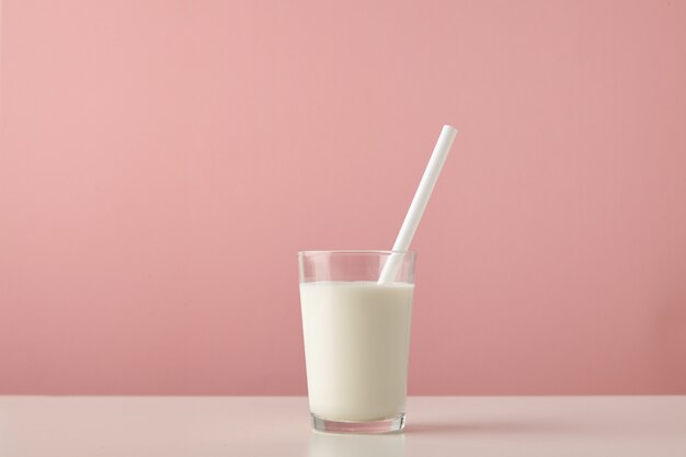 Transparentes Glas mit frischer Bio-Milch und weißem Trinkhalm innen lokalisiert auf pastellrosa Hintergrund auf Holztisch
