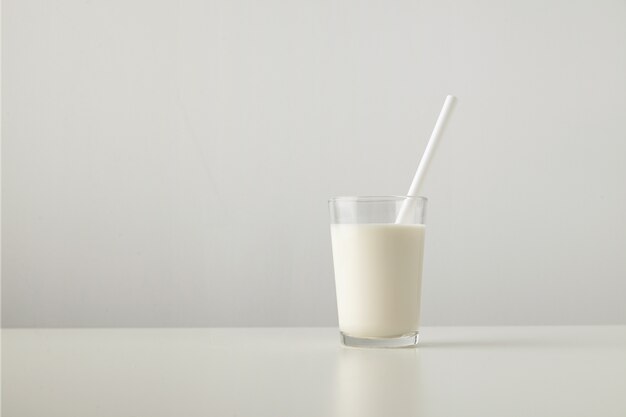 Transparentes Glas mit frischer Bio-Milch und weißem Trinkhalm innen lokalisiert auf Seite des weißen Tisches