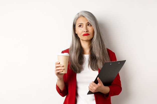Traurige asiatische Geschäftsfrau, die Kaffee bei der Arbeit trinkt und obere linke Ecke schaut, stehend über weißem Hintergrund