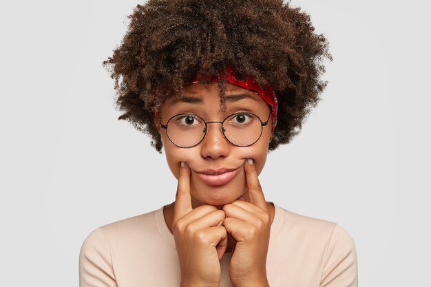 Traurige dunkelhäutige Frau gibt vor zu lächeln, hält beide Zeigefinger in der Nähe der Lippen, bringt ein Lächeln