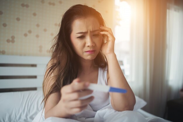 Traurige Frau, die beschwert, einen Schwangerschaftstest sitzend, der auf Bett sitzt