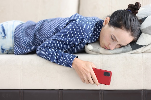Traurige frau liegt auf der couch mit smartphone-anklopfen und einsamkeitskonzept