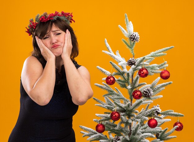 Traurige Frau mittleren Alters, die Weihnachtskopfkranz und Lametta-Girlande um den Hals trägt, die nahe verziertem Weihnachtsbaum stehen und Hände auf Gesicht lokalisiert auf orange Wand hält