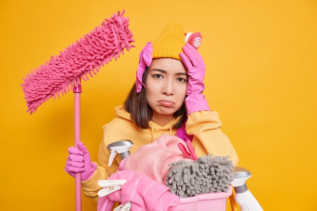 Traurige frustrierte junge Asiatin leidet unter Kopfschmerzen, nachdem sie viel Arbeit über das Haus gemacht hat hasst Wäsche hält Mop, um die Wände des Zimmers einzeln auf gelbem Hintergrund zu reinigen, drückt negative Emotionen aus