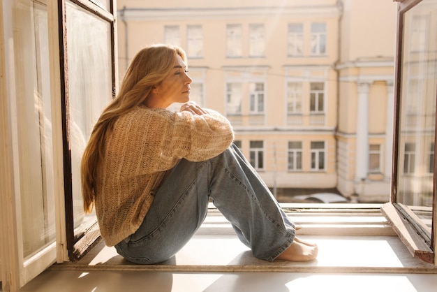 Traurige hellhäutige erwachsene Frau blond in warmem Pullover und Jeans sitzt an einem sonnigen Tag auf der Fensterbank Stimmungskonzept