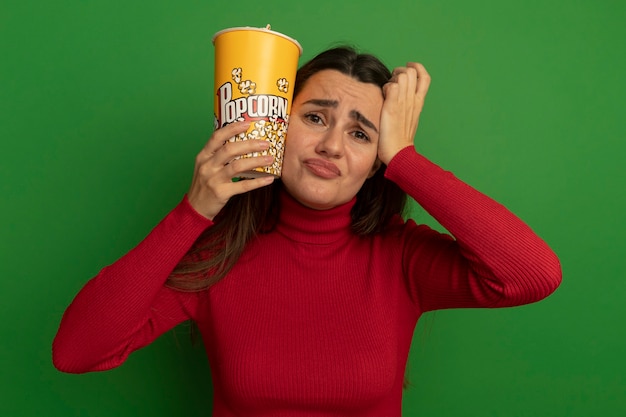 Traurige hübsche Frau hält Eimer Popcorn und legt Hand auf Kopf isoliert auf grüne Wand