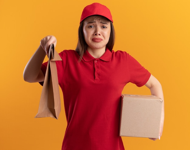 Traurige hübsche Lieferfrau in Uniform hält Papierpaket und Karton auf Orange
