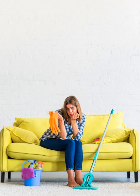 Traurige junge Frau, die auf dem gelben Sofa betrachtet orange Gummihandschuhe sitzt
