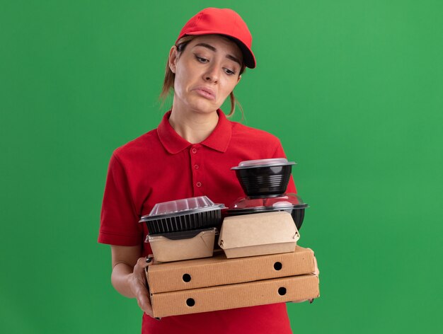 Traurige junge hübsche Lieferfrau in der Uniform hält Papiernahrungsmittelpakete und -behälter auf Pizzaschachteln lokalisiert auf grüner Wand