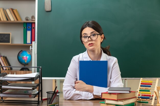 Traurige junge Lehrerin mit Brille, die einen Ordner hält, der am Tisch mit Schulwerkzeugen im Klassenzimmer sitzt