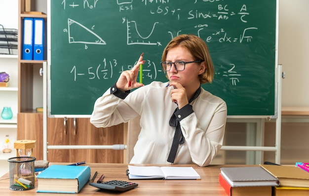 Traurige junge Lehrerin sitzt am Tisch mit Schulmaterial, das einen Bleistift hält und betrachtet das Kinn im Klassenzimmer